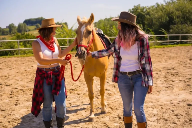 Two women in cowboy hats and plaid shirts interacting with a saddled horse in an outdoor ranch setting.