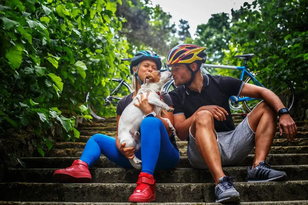 A couple in cycling gear sitting on outdoor steps, cuddling a small dog, with bicycles in the background.